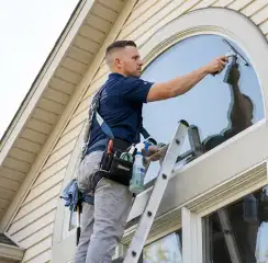 Professional window cleaner on a ladder using a squeegee to clean an exterior arched window, South Bend, IN.
